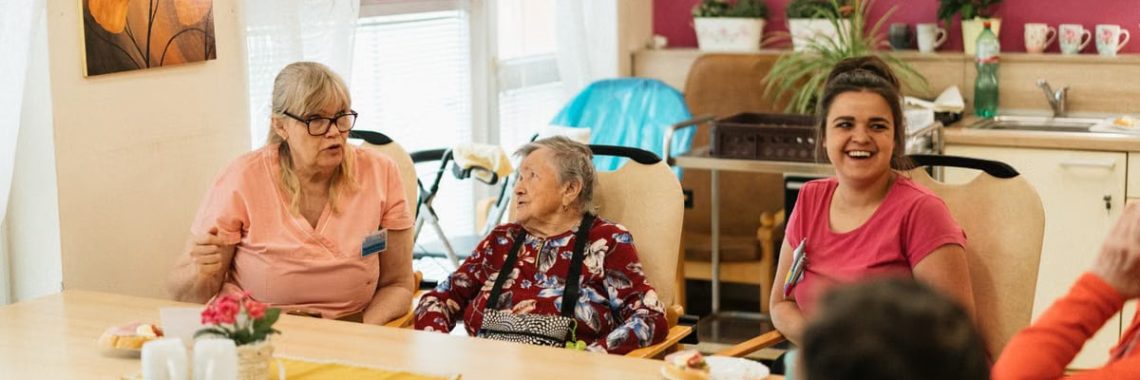 Elderly residents and caregivers enjoying a meal and conversation in a cheerful dining space.